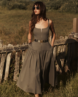 A woman with long brown hair wearing sunglasses and a sleeveless, belted olive-green dress stands by a rustic wooden fence in a grassy outdoor setting.