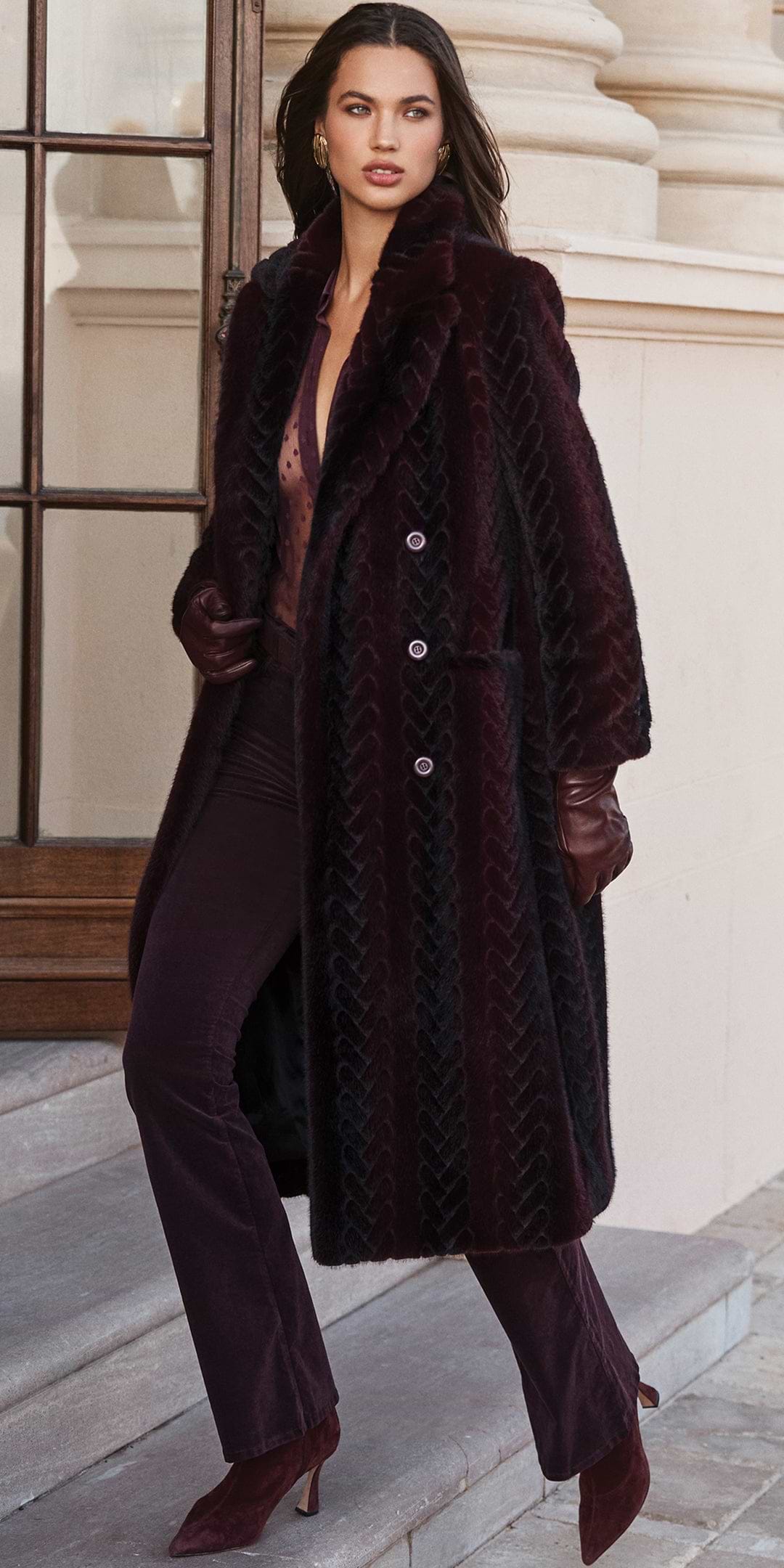 A woman wears a long, deep purple patterned coat over a coordinating shirt and flared trousers, complemented by leather gloves and boots.
