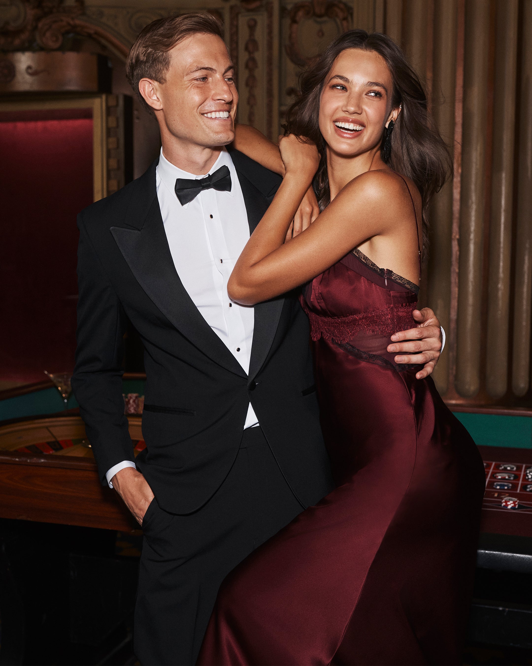 A man in a black tuxedo and a woman in a deep red slip dress posing together in an elegant casino.