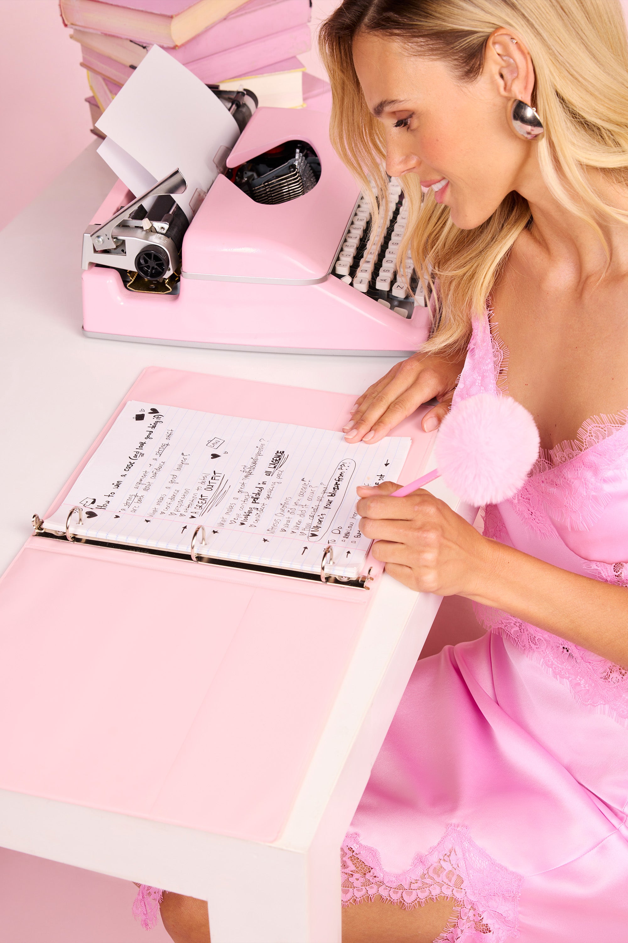 Una donna con un abito di raso rosa è seduta a una scrivania bianca e sta scrivendo su un quaderno con le pagine rosa. Sulla scrivania ci sono una macchina da scrivere rosa e una pila di libri rosa. La scena ha unestetica delicata, dai toni pastello.