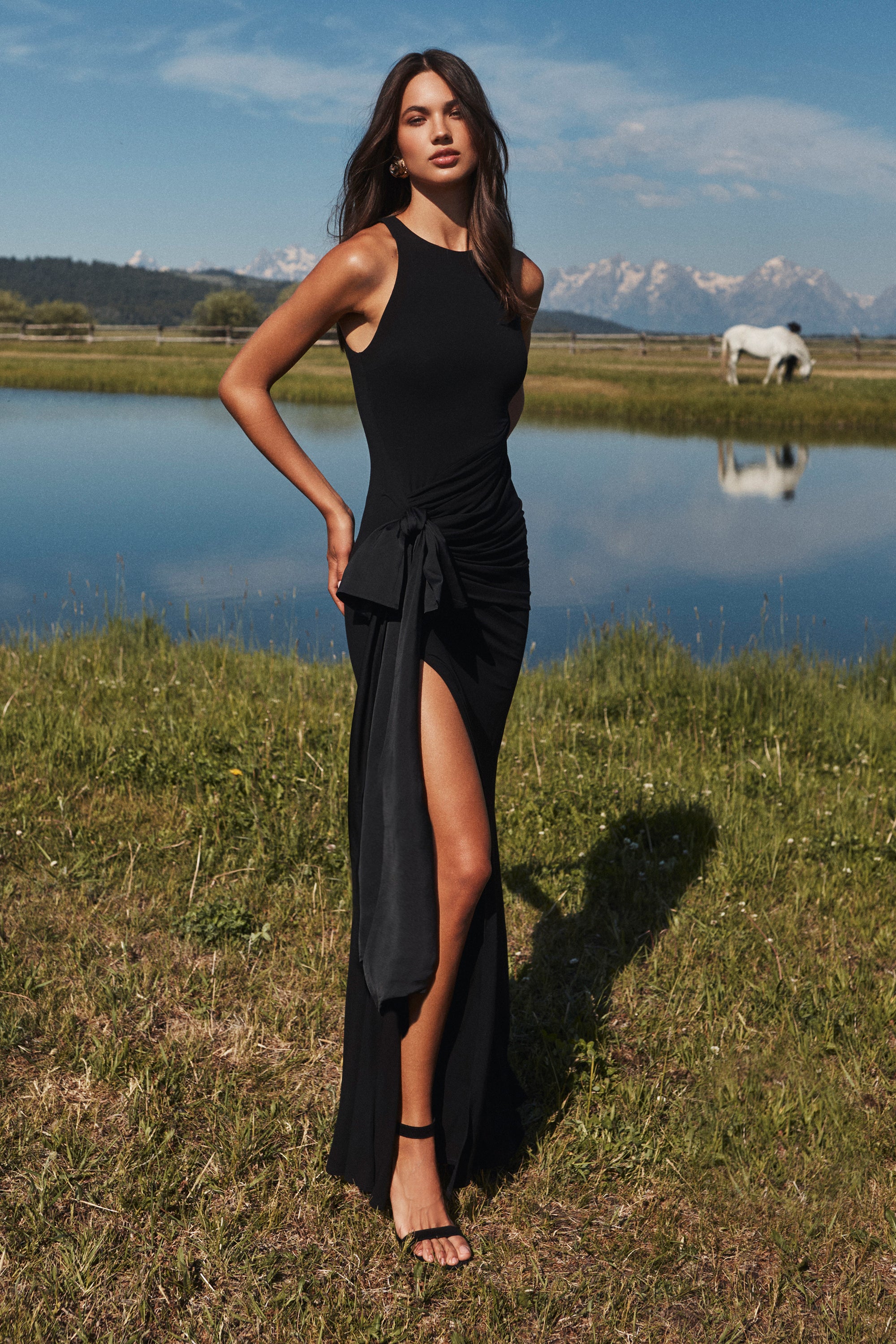 Woman in a sleeveless black dress with a high slit stands on grass near a calm pond, with a white horse grazing in the background under a blue sky.