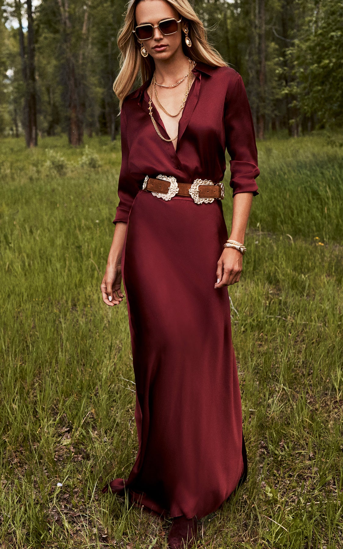 Woman wearing a long, deep red dress with rolled sleeves, a wide decorative belt, sunglasses, and jewelry, standing on grass in a wooded outdoor setting.