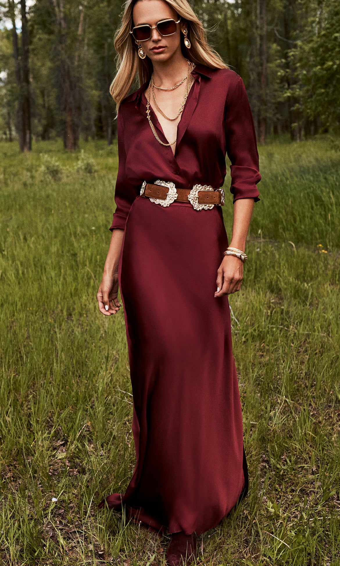 Woman standing on grass wearing a long, burgundy silk dress with rolled sleeves, a wide decorative belt, layered necklaces, sunglasses, and bracelets, with trees in the background.