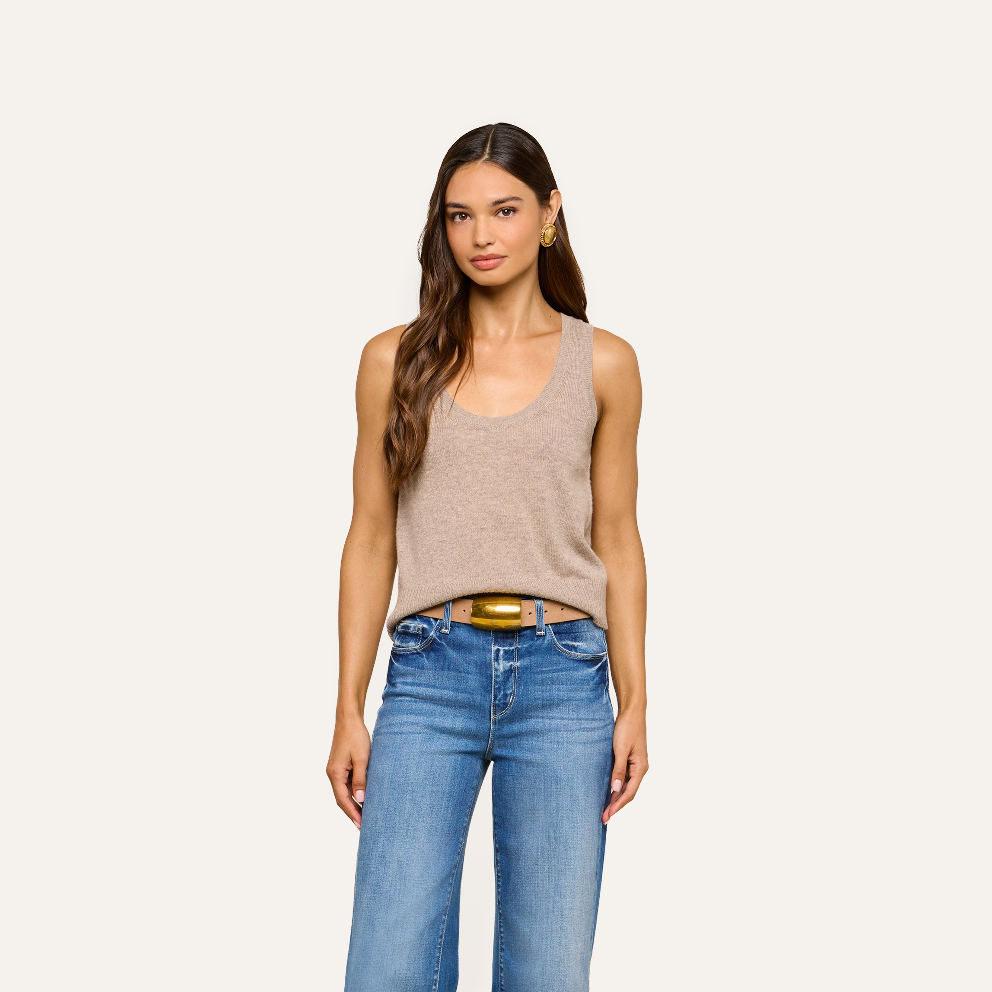 A woman with long brown hair, dressed in a tan tank top, blue jeans, and a brown belt, stands confidently facing forward against a plain light background.