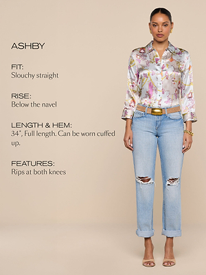 A woman stands facing forward wearing a shiny patterned blouse and light blue slouchy straight jeans with rips at both knees. Text describes jeans: slouchy fit, rise below navel, 34 full length, rips at both knees, can be cuffed.