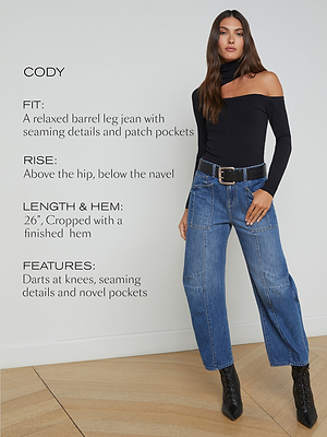 A woman wears a black off-the-shoulder top and relaxed barrel leg jeans with patch pockets and a belt, standing indoors. Text describes the jeans’ fit, rise, length, hem, and features.