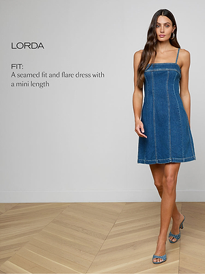 A woman with long brown hair models a sleeveless, blue denim flare mini dress with visible seams. She stands indoors on a wooden floor, wearing blue heeled sandals. Text on the image describes the dress as seamed fit and flare.