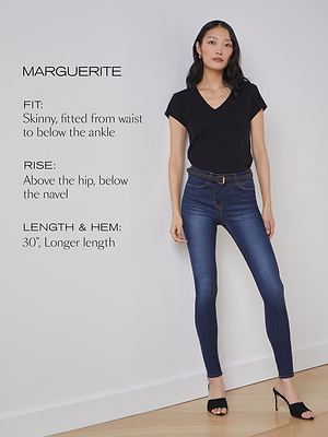 A woman stands against a white wall wearing a black V-neck t-shirt, dark blue skinny jeans, and black heels. Text describes the jeans: skinny fit, rise above the hip and below the navel, 30-inch longer hem.