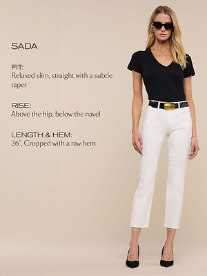 A woman wearing black sunglasses, a black V-neck T-shirt, white cropped slim pants with a raw hem, a black belt with a gold buckle, and black heels stands against a beige background. Text describes the pant fit, rise, and length.