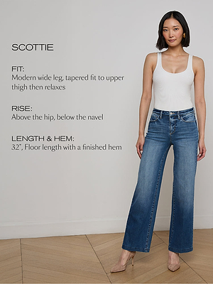 A woman wearing a white tank top and blue wide-leg jeans stands on a wooden floor. Text details the jeans: FIT: Modern wide leg, tapered fit to upper thigh then relaxes. RISE: Above the hip, below the navel. LENGTH & HEM: 32, floor length, finished hem.
