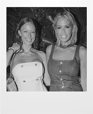 Two women posing together and smiling at the camera; one wears a strapless white dress, the other wears a sleeveless buttoned denim dress. The background features leafy plants. The photo is in black and white.