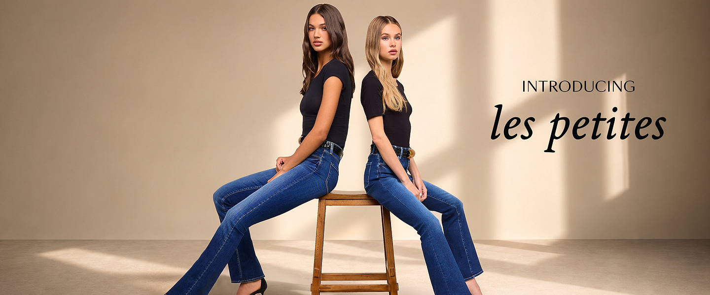 Two women wearing black tops and blue jeans sit back-to-back on a wooden stool. Text beside them reads INTRODUCING les petites, set against a neutral beige background with soft lighting.