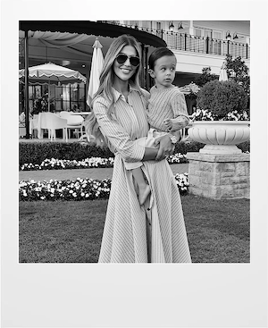 A woman wearing sunglasses and a long striped dress smiles while holding a young child outside in a garden with flowers, patio tables, and umbrellas in the background.