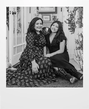 Two women sit side by side indoors, smiling at the camera. Both wear long dark dresses, and one has cowboy boots. They are surrounded by plants and framed art in a cozy room. The photo is in black and white.