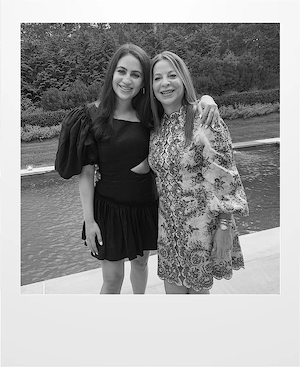Two women stand side by side, smiling with arms around each other, in front of a pool and garden. One wears a black dress with puffed sleeves; the other wears a patterned dress with long sleeves. The image is in black and white.