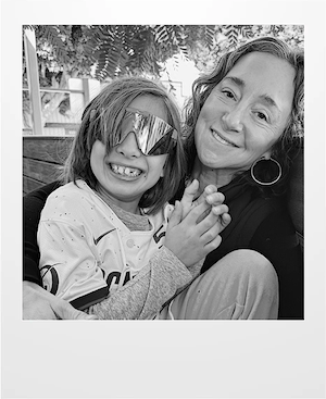 A smiling child wearing large reflective sunglasses and a sports jersey sits on a woman’s lap. The woman, also smiling, has wavy hair and hoop earrings. They are holding hands and sitting closely outdoors.