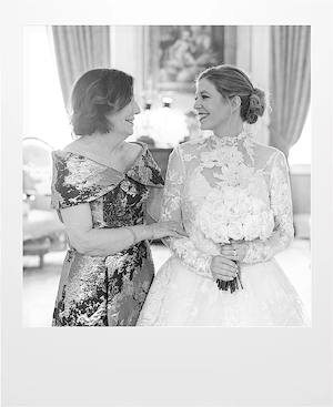 A bride in a lace wedding dress holds a bouquet of roses and smiles at a woman in a floral off-the-shoulder gown; they stand close together in an elegant room with large windows and drapes.