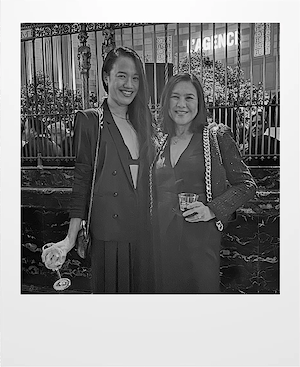 Two women dressed in formal black outfits stand side by side, smiling at the camera. One holds a wine glass and the other a drink, with a decorative fence and city lights behind them. The image is in black and white.
