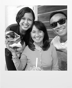 Three people smile at the camera; a woman on the left holds a small dog, a woman in the center is sitting with a drink, and a man on the right wears sunglasses. The photo is in black and white.