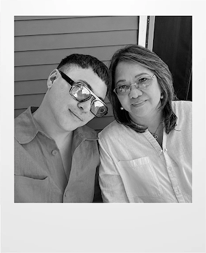 A young man wearing sunglasses and a button-up shirt sits next to an older woman with glasses and shoulder-length hair. Both are smiling slightly and posing for a black-and-white photo in front of a clapboard wall.