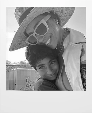 A woman wearing a wide-brimmed hat and large sunglasses smiles as she hugs a young child who is smiling at the camera. The photo is in black and white and taken outdoors.