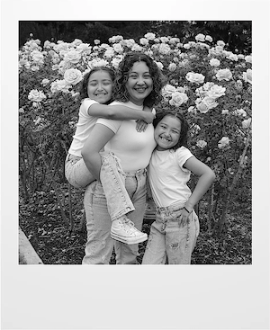 Three people smile in front of blooming rose bushes. One person stands in the center holding a child piggyback style, while another child stands beside them, all wearing jeans and white tops.