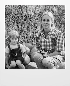 A black-and-white photo of a woman in a checkered shirt and a young girl in overalls sitting together outdoors, surrounded by pumpkins and tall grass. Both are looking at the camera and smiling gently.