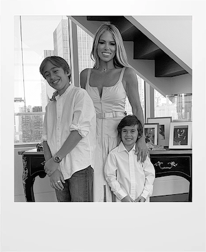 A woman stands smiling with two young boys in front of a large window, all dressed in white shirts. They are indoors, next to a dark table with framed photos, and a staircase is visible behind them.
