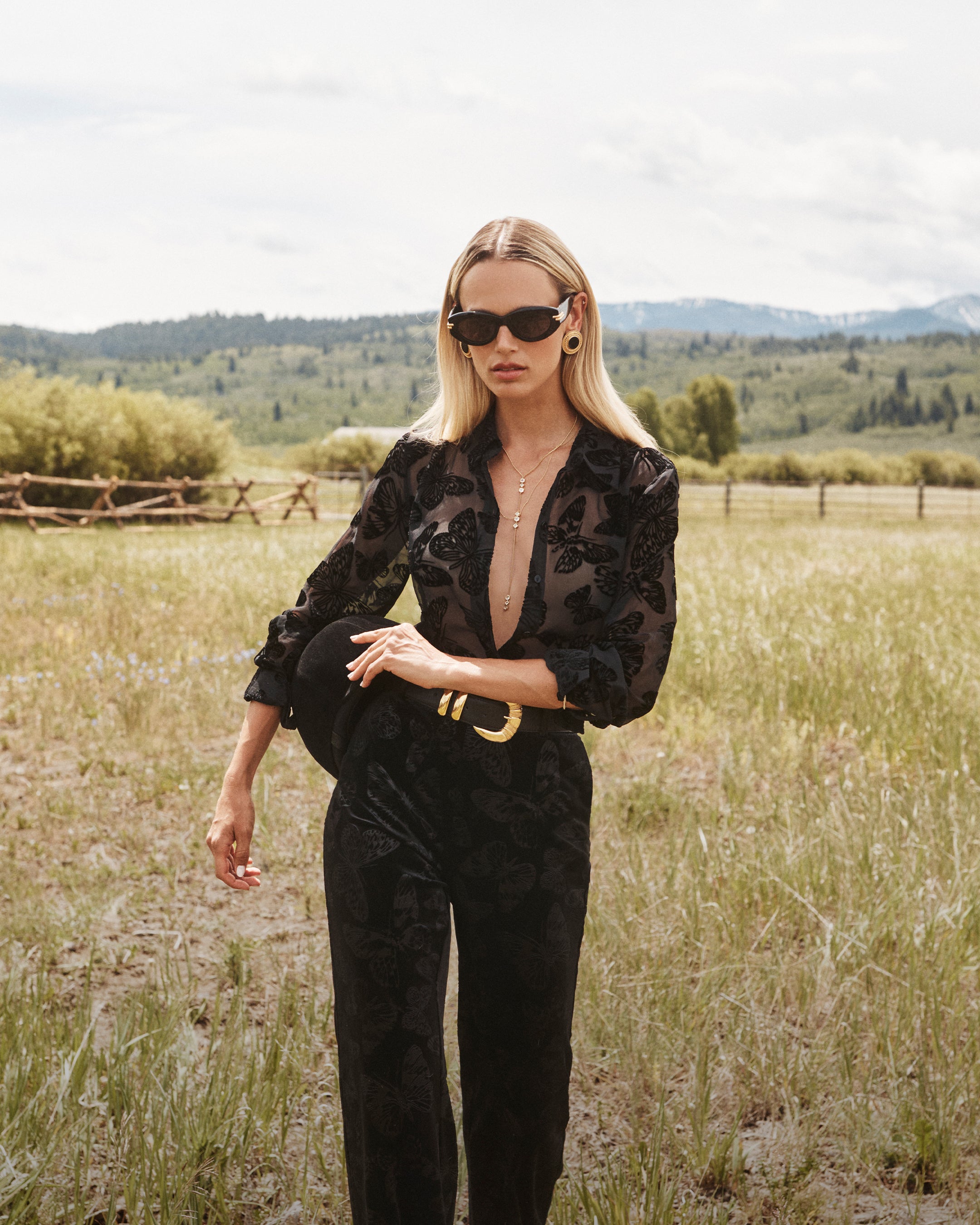 Woman in sunglasses wearing a sheer black patterned blouse and black pants stands in a grassy field with mountains and trees in the background, holding a black hat.
