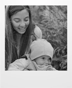 A woman smiles while leaning close to a young child wearing a beanie and a jacket, both outdoors surrounded by blurred foliage in the background. The image is in black and white.