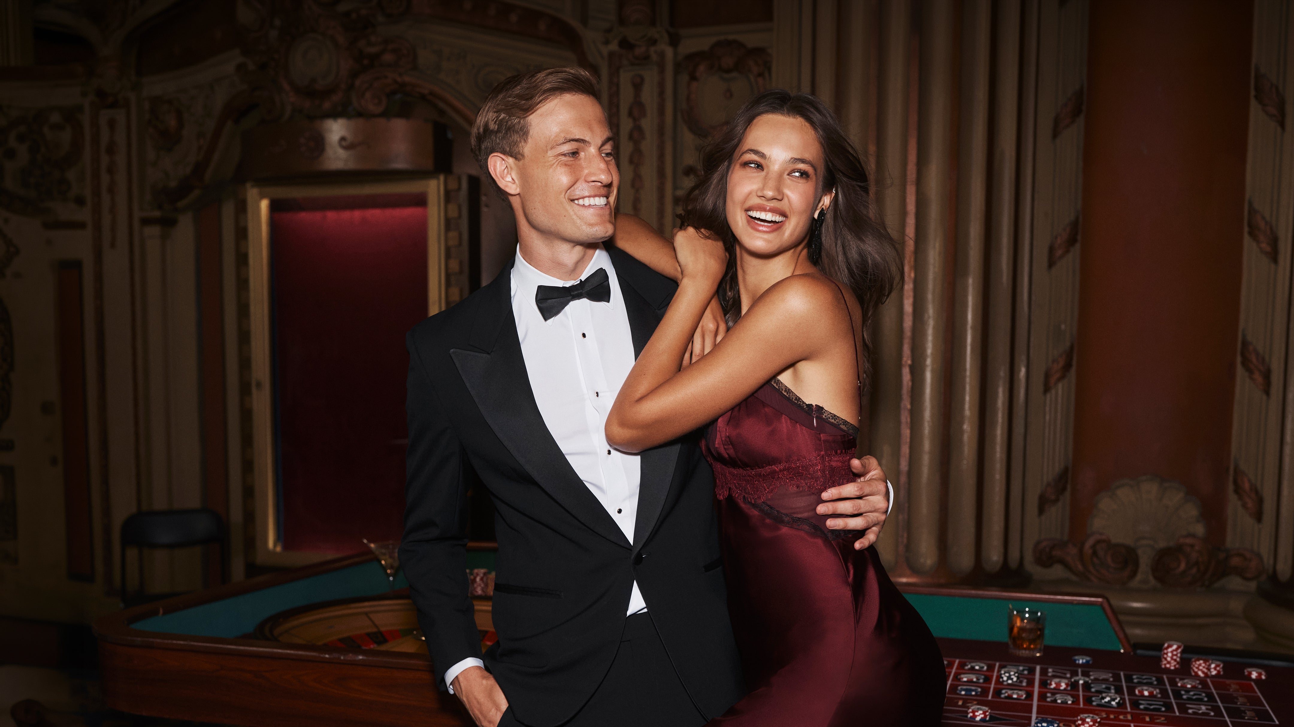 A man in a tuxedo and a woman in a dark red slip dress laughing together at a roulette table in a dimly lit, ornate casino room.