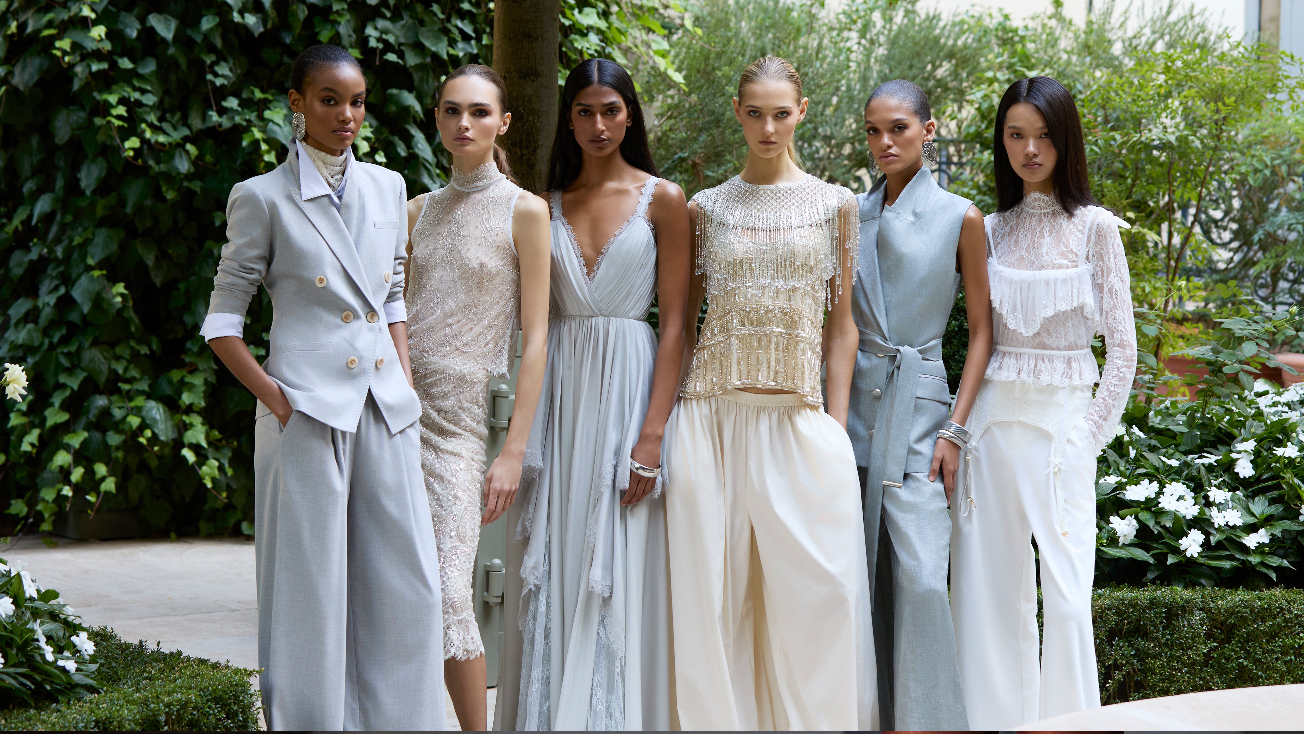 Six models stand side-by-side in a garden, wearing neutral-toned formal outfits including suits, dresses, and embellished tops.