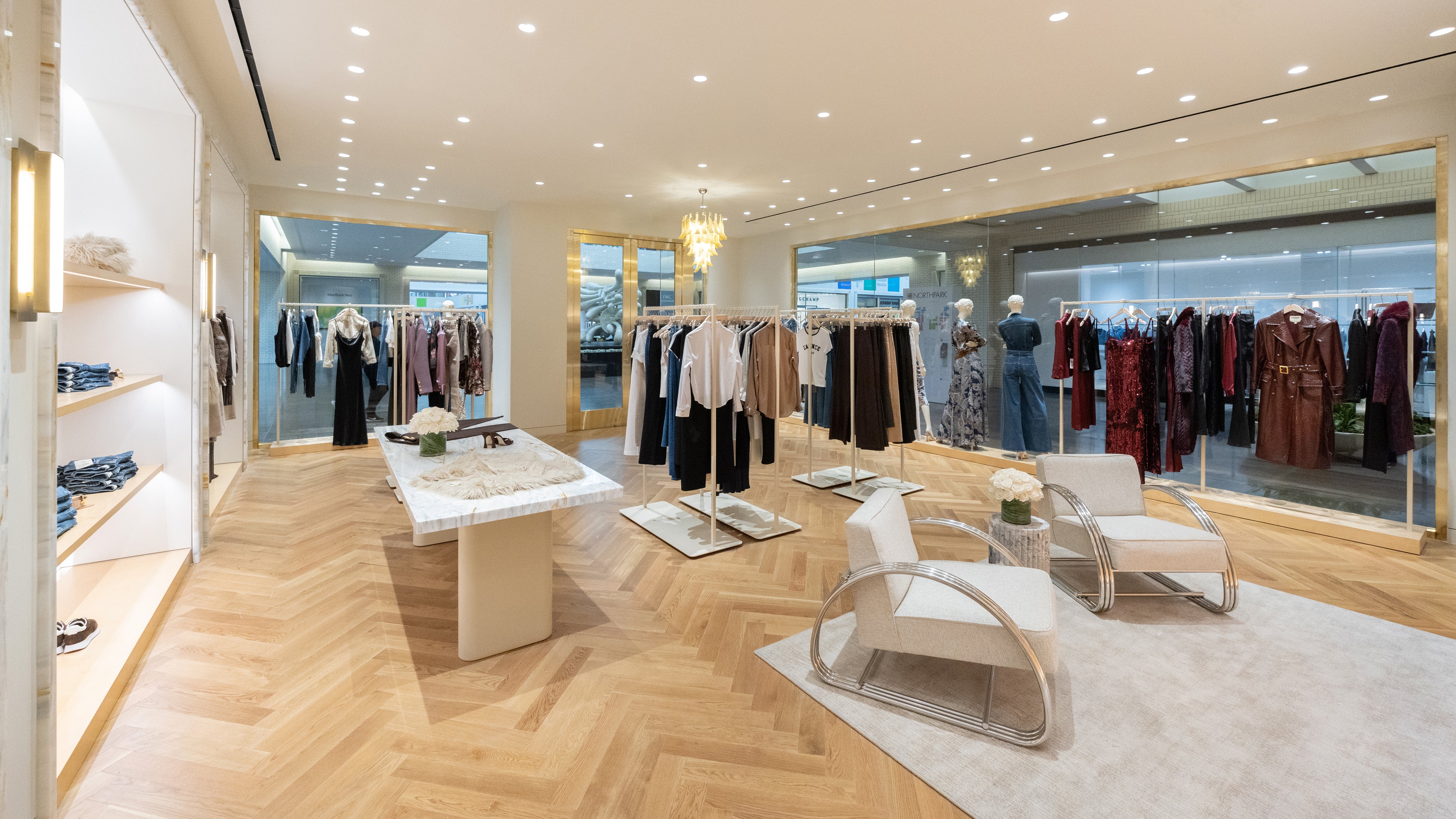 A boutique interior featuring racks of hanging garments, a marble display table, and two modern lounge chairs arranged on a light-colored rug.