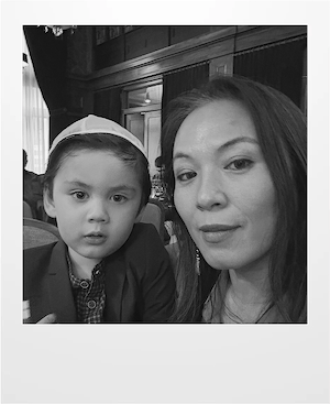 A black-and-white photo of a woman and a young boy sitting indoors, both looking at the camera. The boy is wearing a blazer and a cap, and the woman has long hair. The background shows curtains and windows.