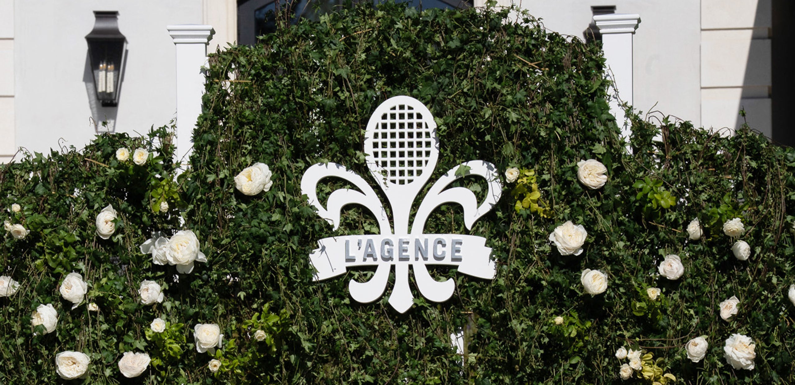 A white logo with a tennis racket and the text LAGENCE is displayed on a green leafy wall decorated with white roses.