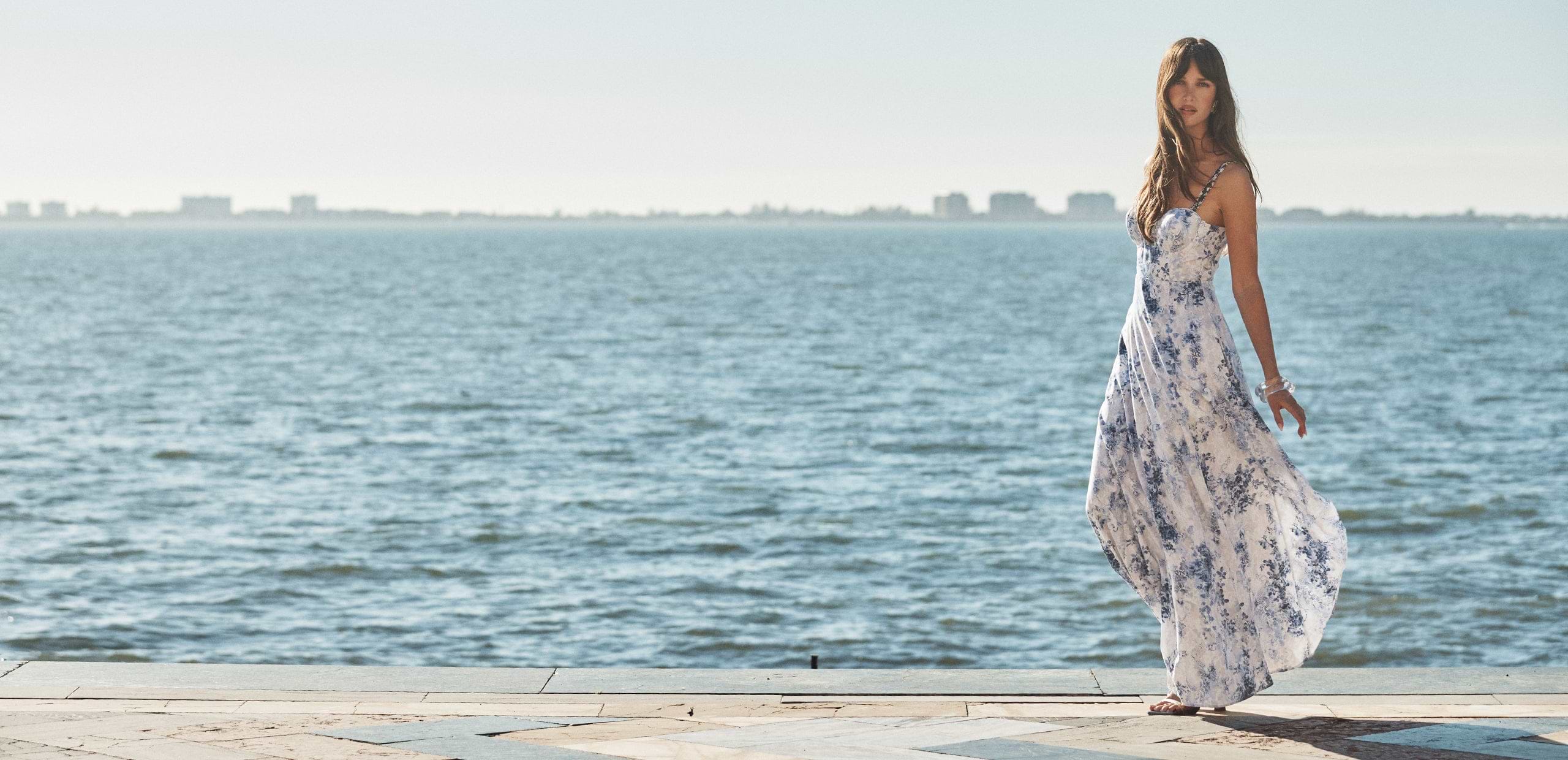 A woman in a flowing white and blue dress stands near the edge of a waterfront, with the sea and distant city buildings in the background on a sunny day.