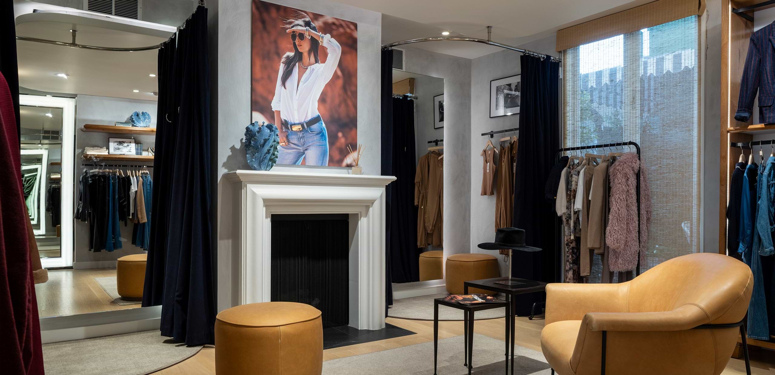 A stylish boutique fitting room with tan leather chairs, clothing racks, mirrors, a white fireplace, and a large fashion portrait above the mantel. Warm lighting and modern decor create a cozy, inviting atmosphere.
