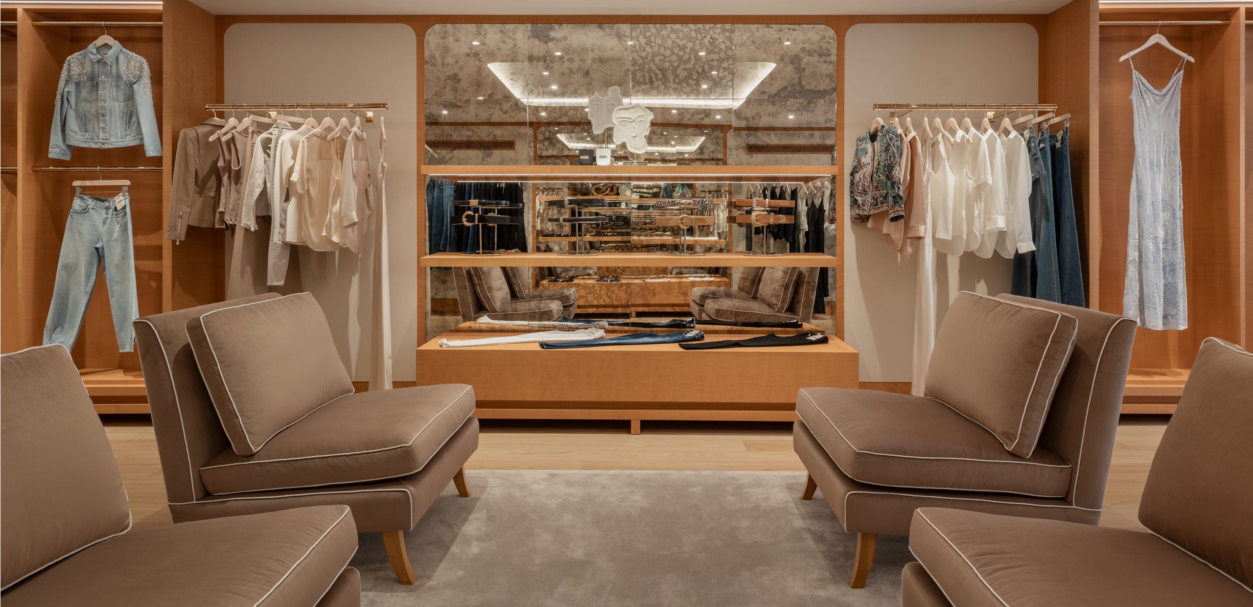 A stylish clothing boutique features neatly hung clothes, a mirrored wall with shelves displaying accessories, and four cushioned chairs arranged on a gray rug, creating a modern and inviting seating area.