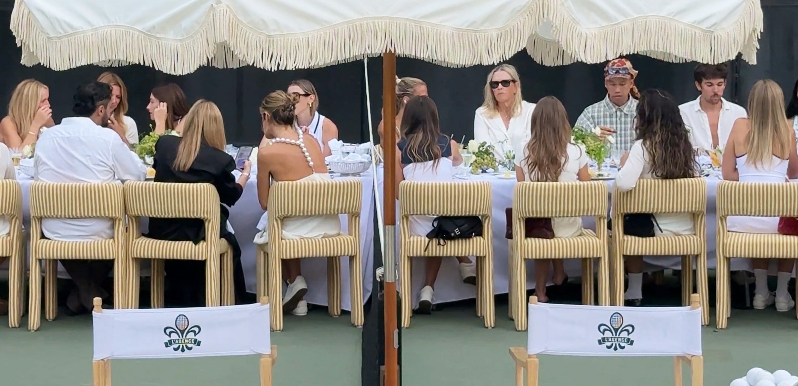 A group of people dressed mostly in white sit around a long, elegant outdoor table under a large white umbrella. Two empty chairs with tennis-themed logos are in the foreground.