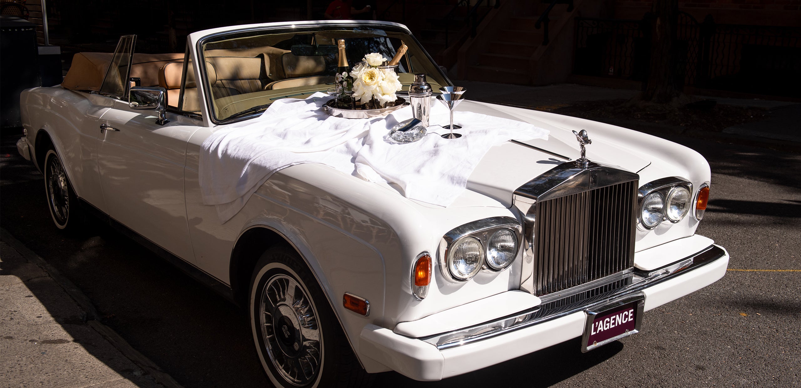 A stylish white convertible adorned with a bouquet of white flowers rests on a city street, its hood elegantly set with two wine glasses and a tray draped in a crisp white cloth.