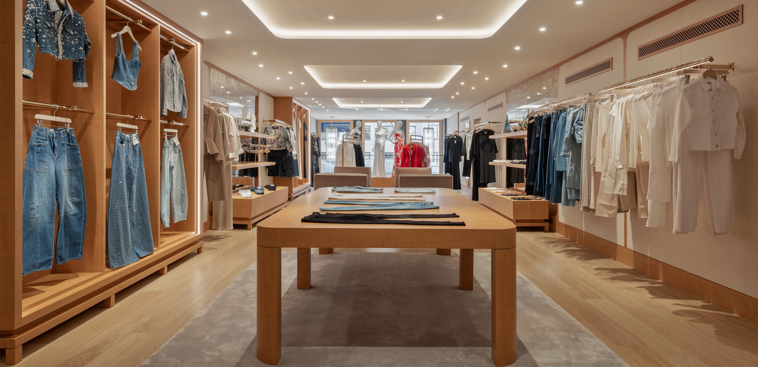 A modern clothing boutique with wooden shelves displaying denim, shirts, and jackets. A central wooden table holds folded clothes, and natural light enters from windows at the far end of the store.