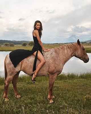 A woman in a black dress and heels sits side-saddle on a brown horse near a lake, with grass, water, and mountains in the background under a cloudy sky.
