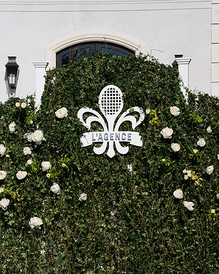 A green wall with white roses displays a white emblem featuring a tennis racket and the text LAGENCE in front of a building.