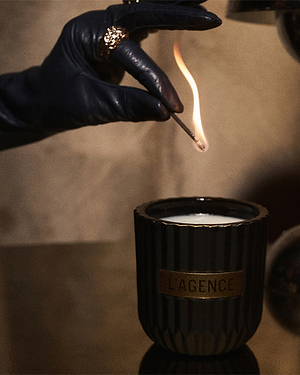 A gloved hand with a ring lights a match above a black, ribbed candle jar labeled LAGENCE on a reflective surface.