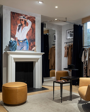 A stylish boutique interior with tan ottomans, a white fireplace, clothing racks, and a large photo of a woman in a white shirt and sunglasses above the mantle. Black curtains and tables with a hat and magazines are visible.