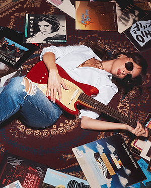 Person playing a red electric guitar surrounded by music albums on a patterned rug