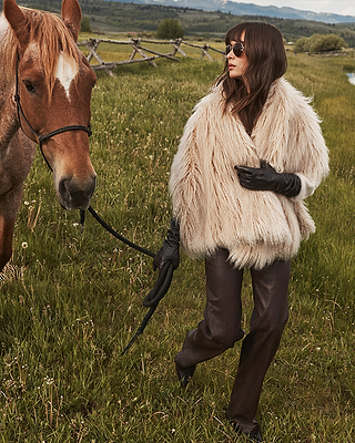 A woman in sunglasses, a fluffy beige coat, brown pants, and black gloves walks through grassy field holding a brown horses reins. A wooden fence and distant hills appear in the background.