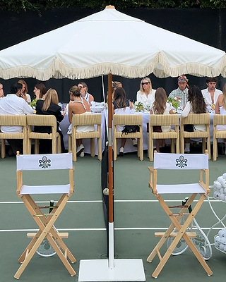 A group of people dressed in white sit at a long table outdoors under a large umbrella, with two empty directors chairs in the foreground on a tennis court.