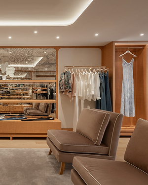 A stylish walk-in closet with beige seating, wood accents, a mirrored wall, clothes hanging neatly on racks, and a light blue dress displayed in a lit wooden alcove.