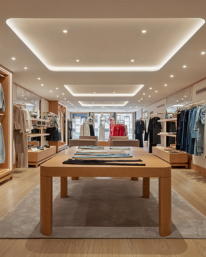 A modern clothing boutique with wooden tables and racks displaying neatly arranged apparel. Soft ceiling lights brighten the space, which features light wood floors and a minimalist, organized layout.